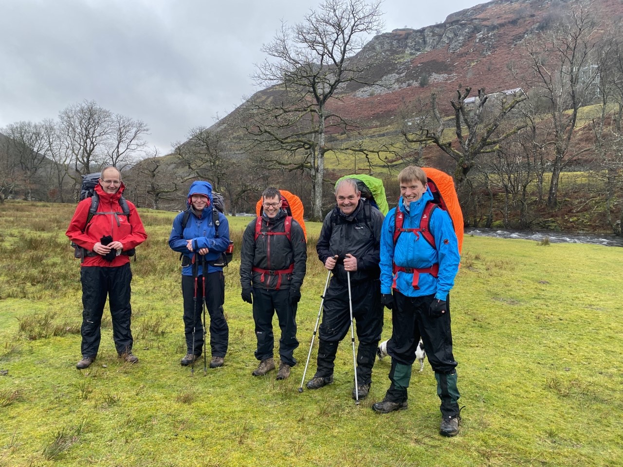 Five happy staff in brightly coloured wet weather gear and large backpacks are ready to trek the mountain behind them in the Elan Valley
